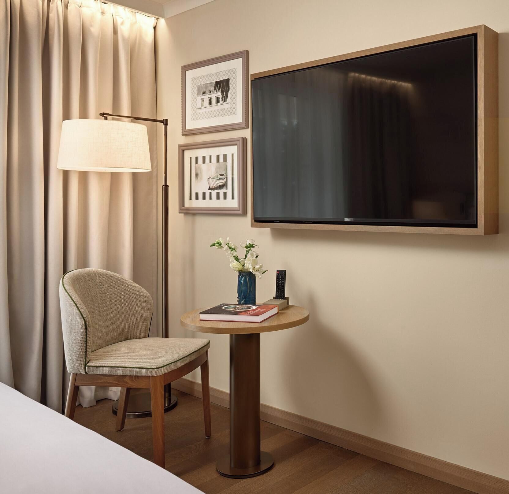 Una camera d'albergo con un letto, una sedia, un tavolino rotondo con un vaso di fiori, una lampada e una TV a parete.