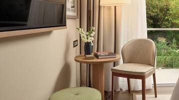 A room with a television mounted on the wall, a round table with a lamp, a chair, and a small ottoman.
