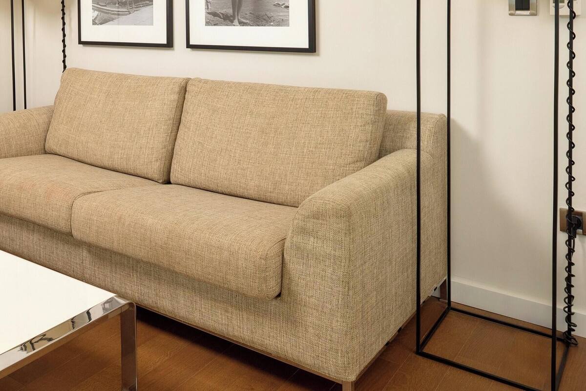 Ein beiges Sofa, ein weißer Tisch mit einer Glasvase und ein Bett im Hintergrund.