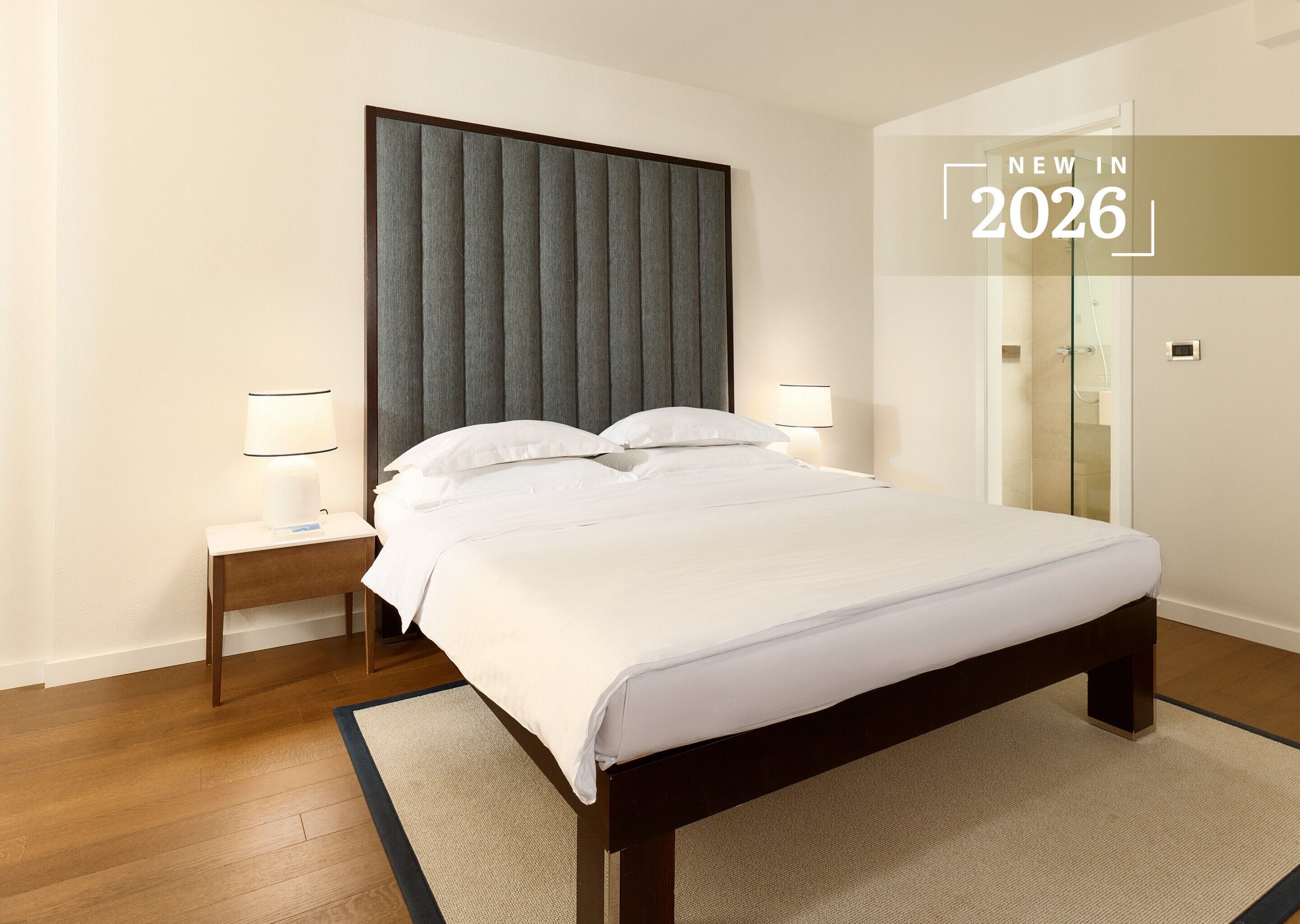 A modern hotel room with a large bed, two bedside tables with lamps, a large window, and a shower area.