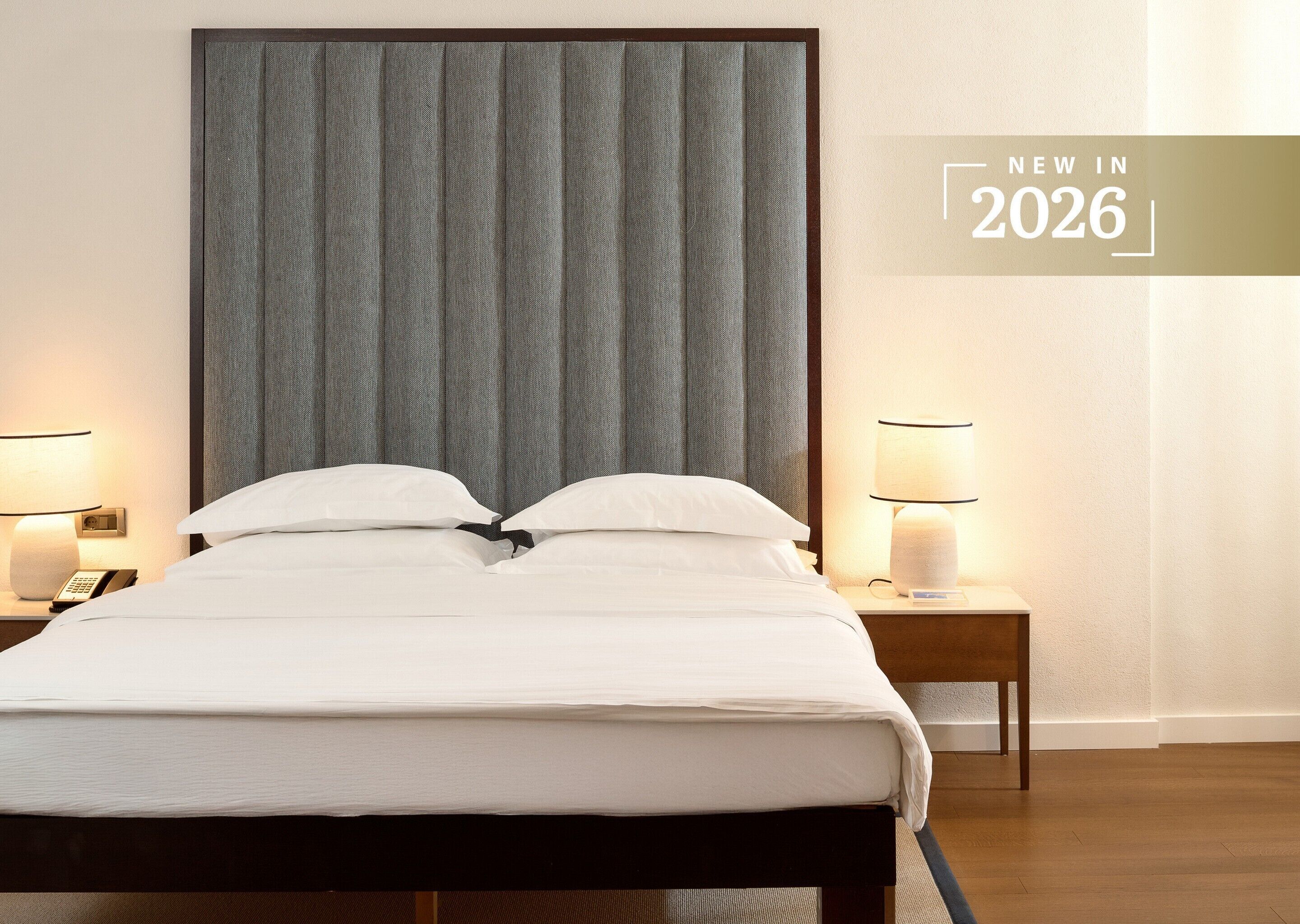 A hotel room with a large bed, two bedside tables with lamps, a telephone, and a wall with a 'NEW IN 2026' sign.