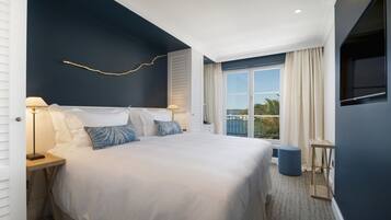 A modern hotel room with a large bed, two bedside tables with lamps, a small stool, a TV mounted on the wall, and a balcony with curtains.