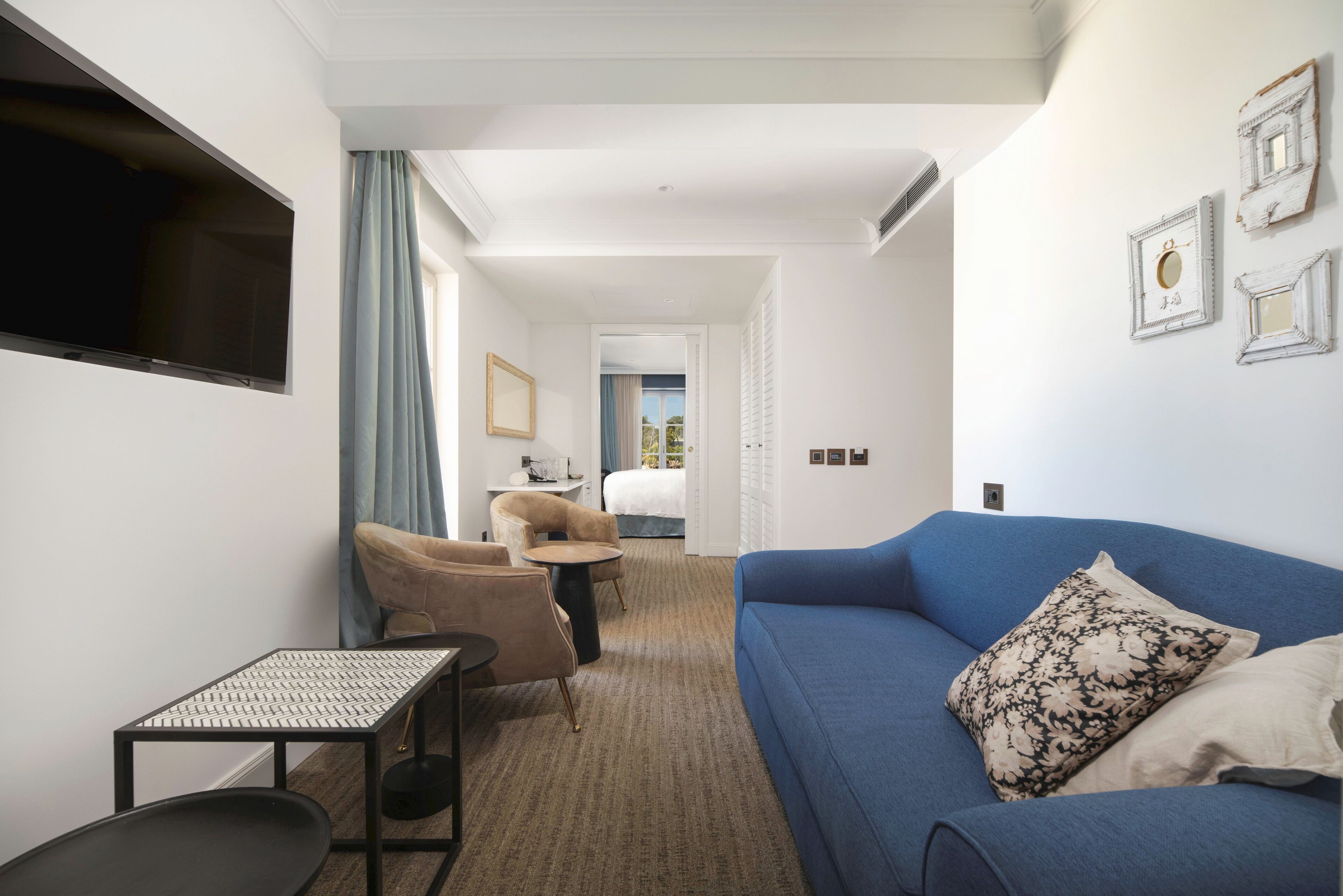 Habitación de hotel con un sofá azul, una mesita pequeña, una silla y un televisor montado en la pared.