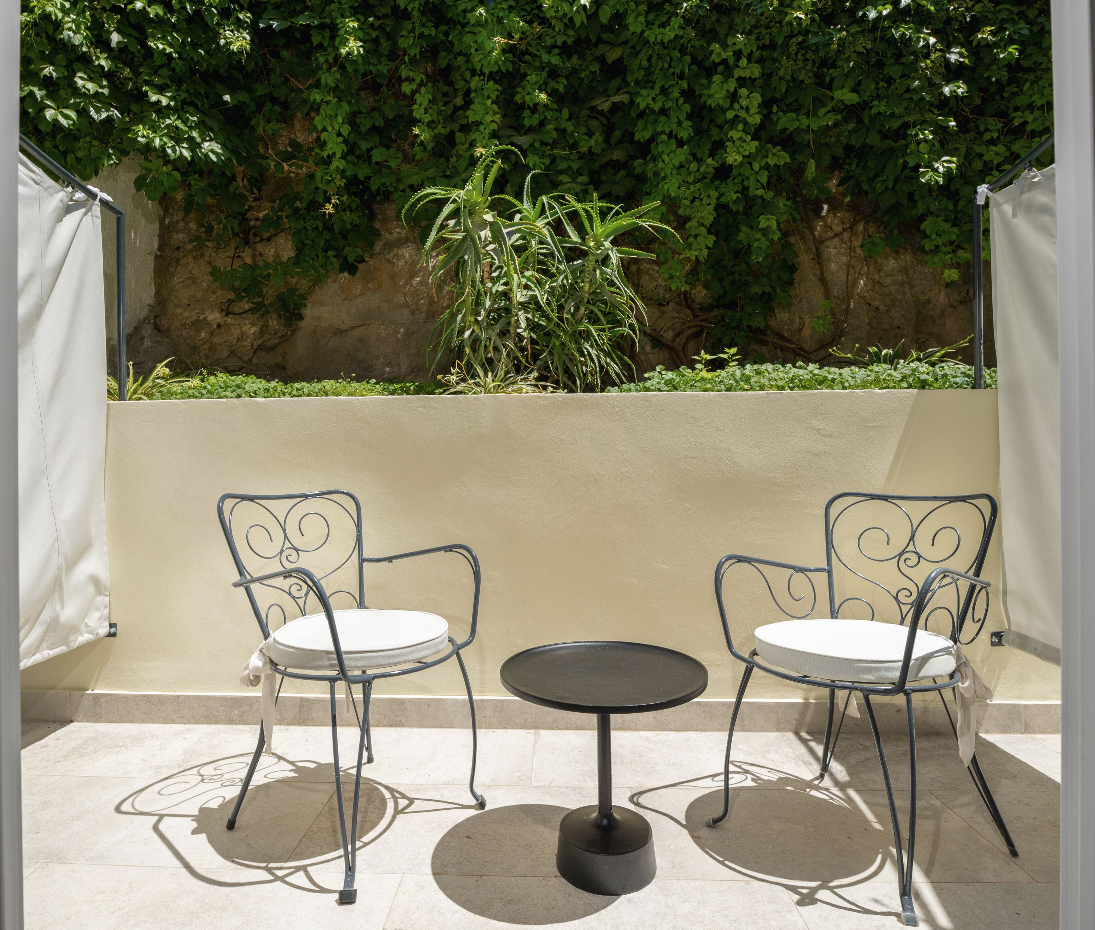 A patio with two metal chairs and a small round table, surrounded by a beige wall and lush green foliage.