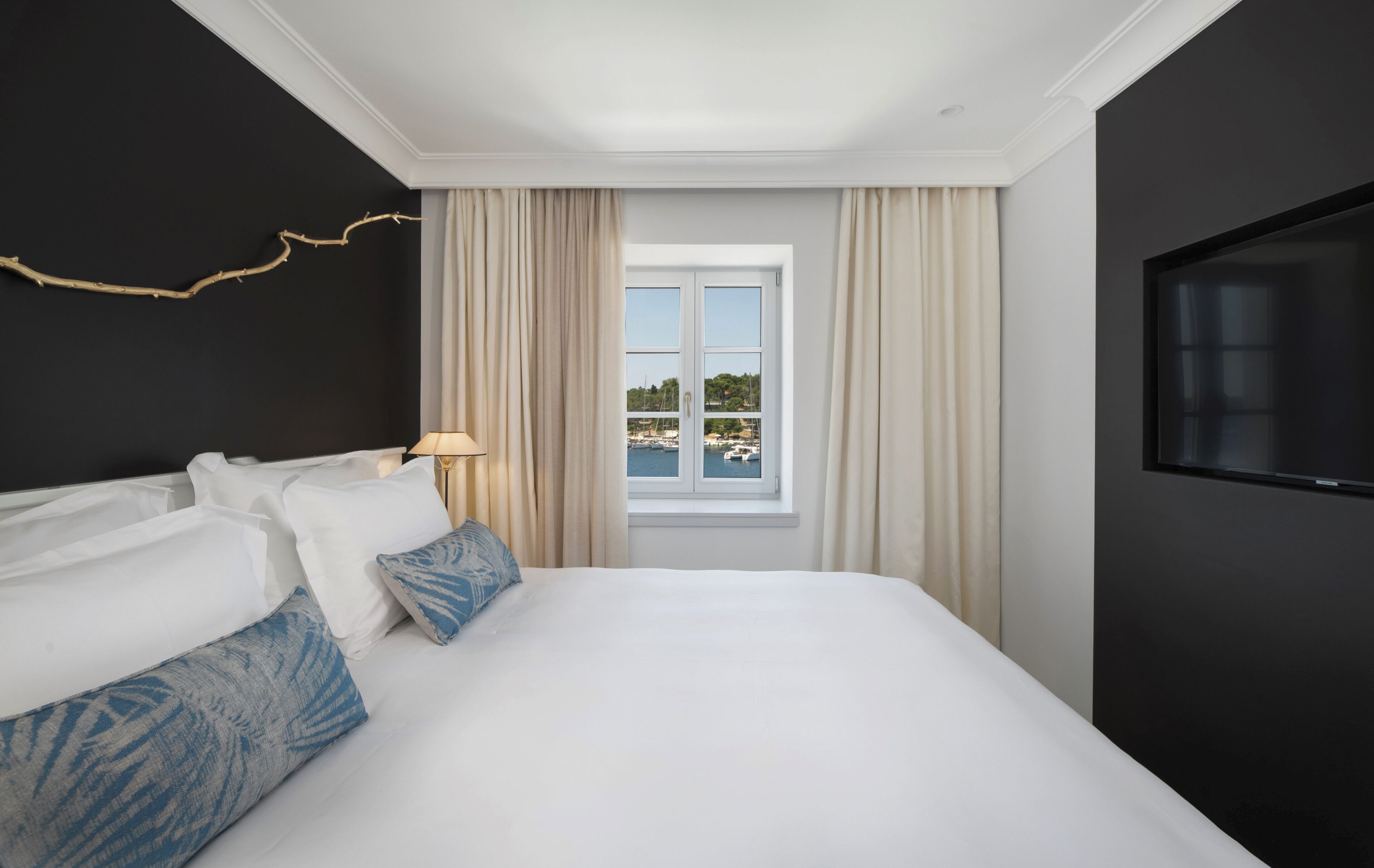 A hotel room with a large bed, a TV, a window with a view of boats, and a decorative branch on the wall.