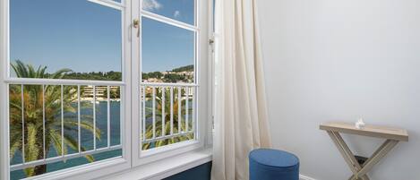 A room with a large window offering a view of a coastal area, a blue ottoman, and a small wooden side table.