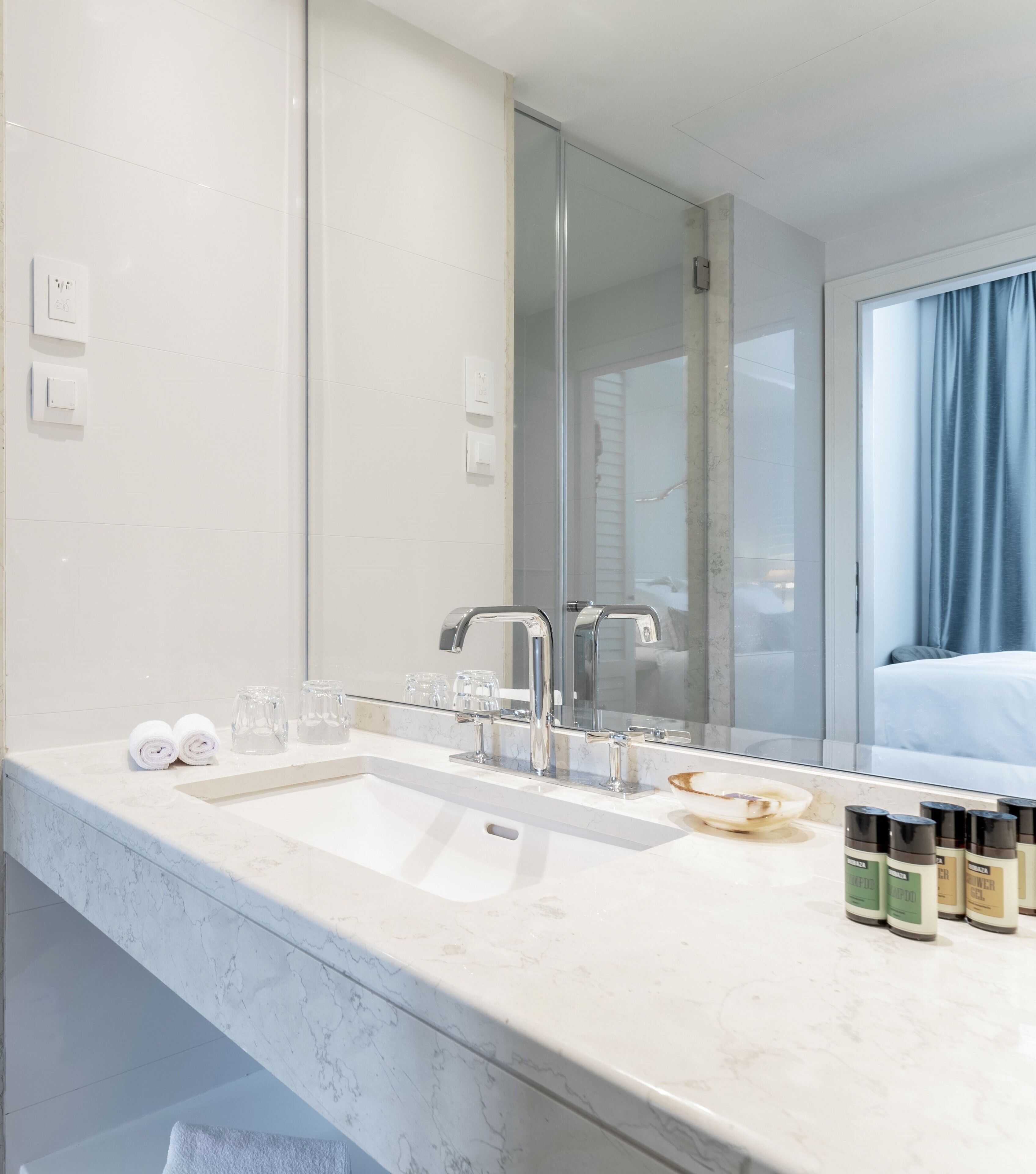 A modern bathroom with a large mirror, a sink, and a vanity. There are toiletries on the counter and a towel rolled up on the counter.