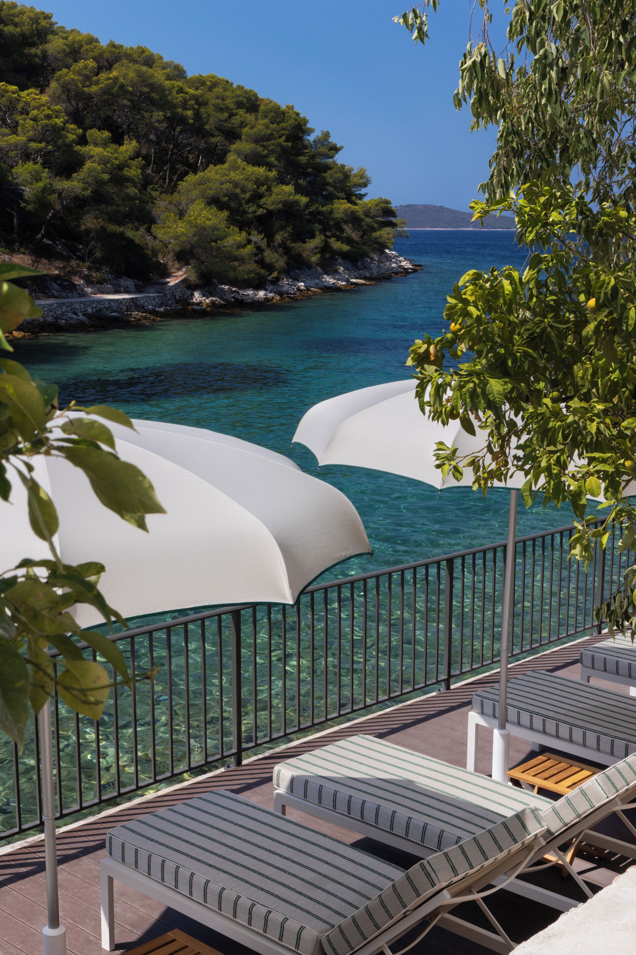 A seaside resort with lounge chairs and umbrellas overlooking a clear blue sea and a forested hillside.