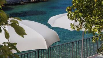 A seaside resort with lounge chairs and umbrellas overlooking a clear blue sea and a forested hillside.