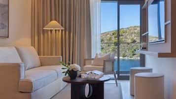 Una habitación de hotel moderna con un sofá, una mesa de centro con un florero y una silla, y vistas a un paisaje costero a través de amplias ventanas.