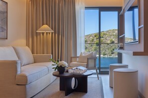 A modern hotel room with a sofa, a coffee table with a vase of flowers, a chair, and a view of a coastal landscape through large windows. - Sirena Resort (Hvar)