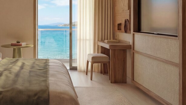 Una habitación de hotel con una cama grande, una pequeña mesa con libros, un escritorio con una silla y un televisor. La habitación tiene vistas al mar y a un paisaje montañoso.