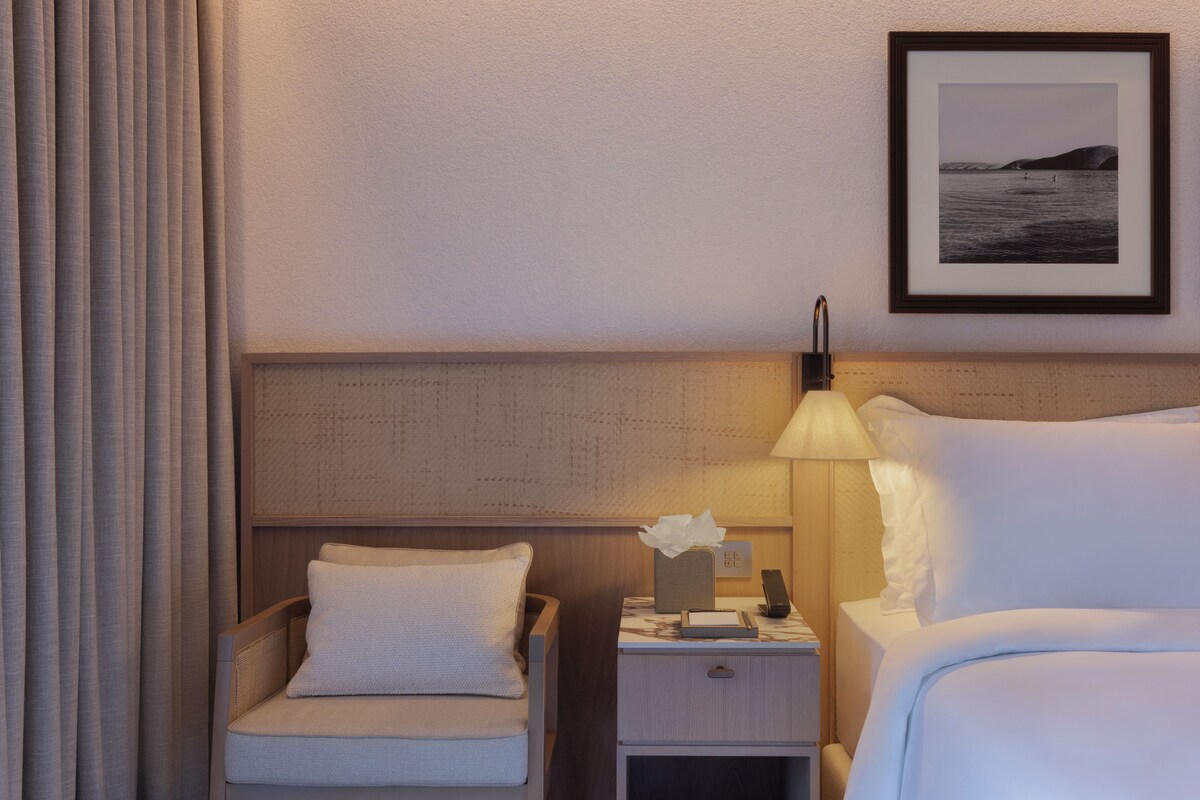 A hotel room with a bed, a nightstand with a lamp, a chair, and a framed picture on the wall.