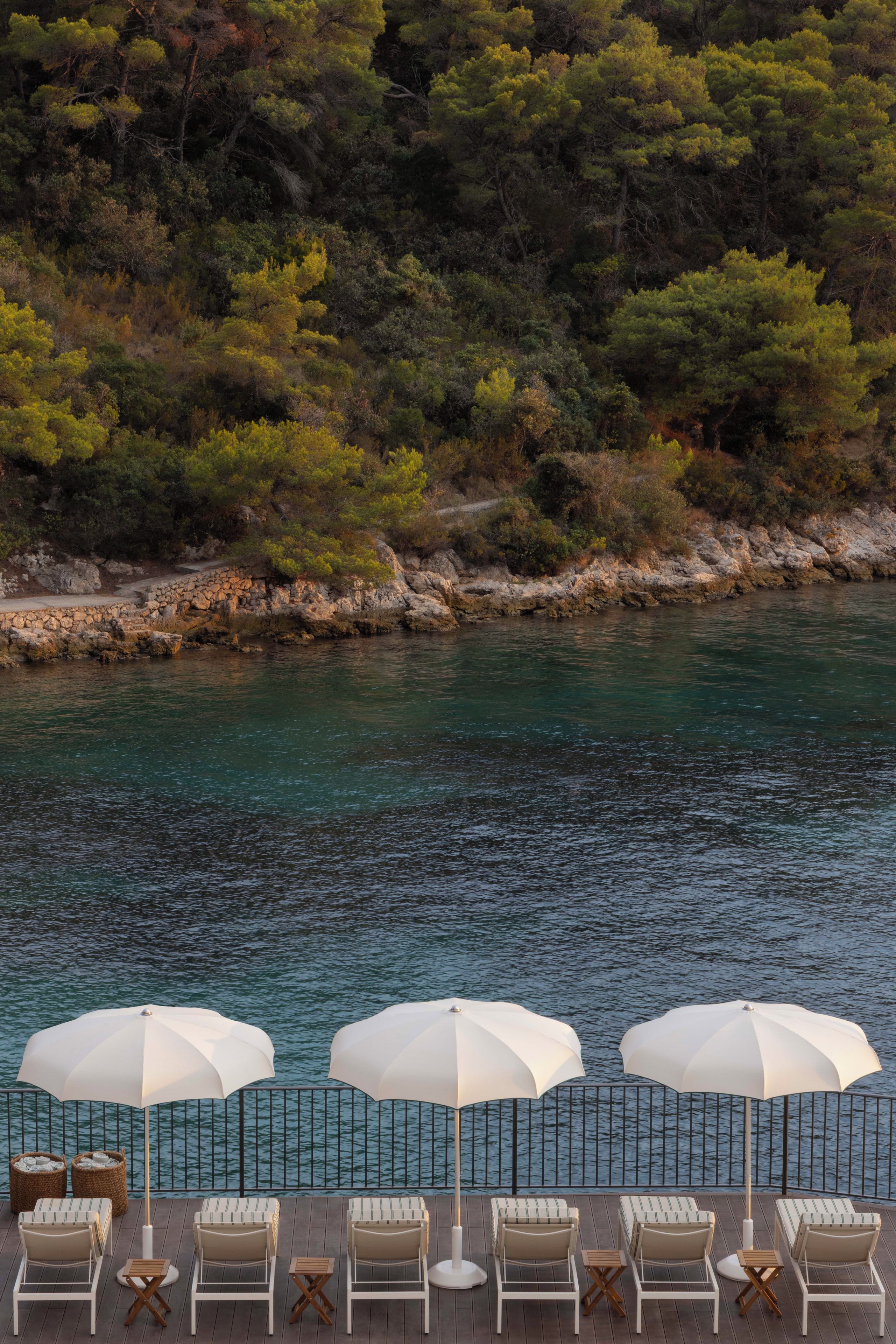 A poolside area with lounge chairs and umbrellas, overlooking a body of water and a wooded hillside.
