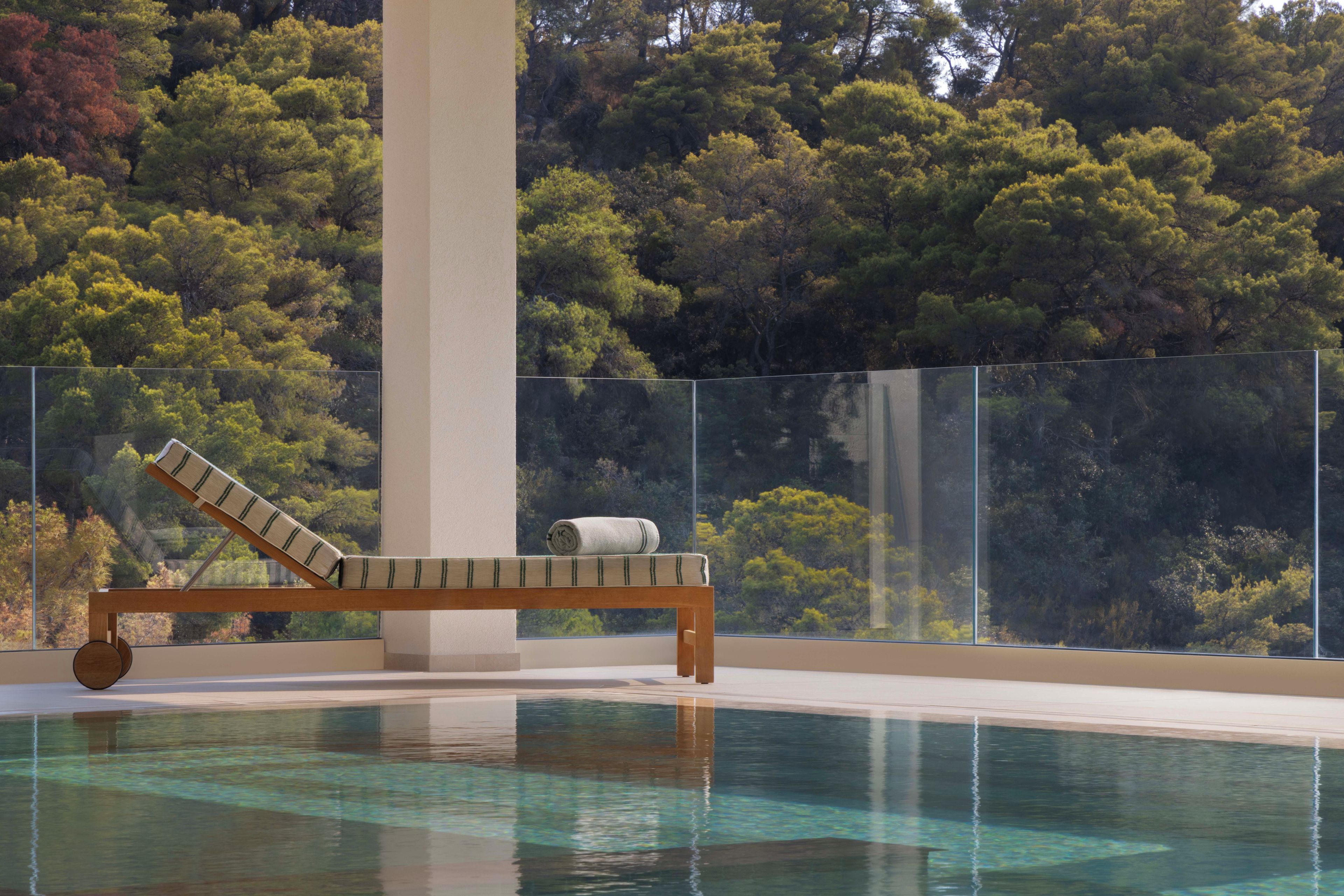 A pool with a wooden lounge chair and a towel, surrounded by a glass barrier, with a forested hillside in the background.