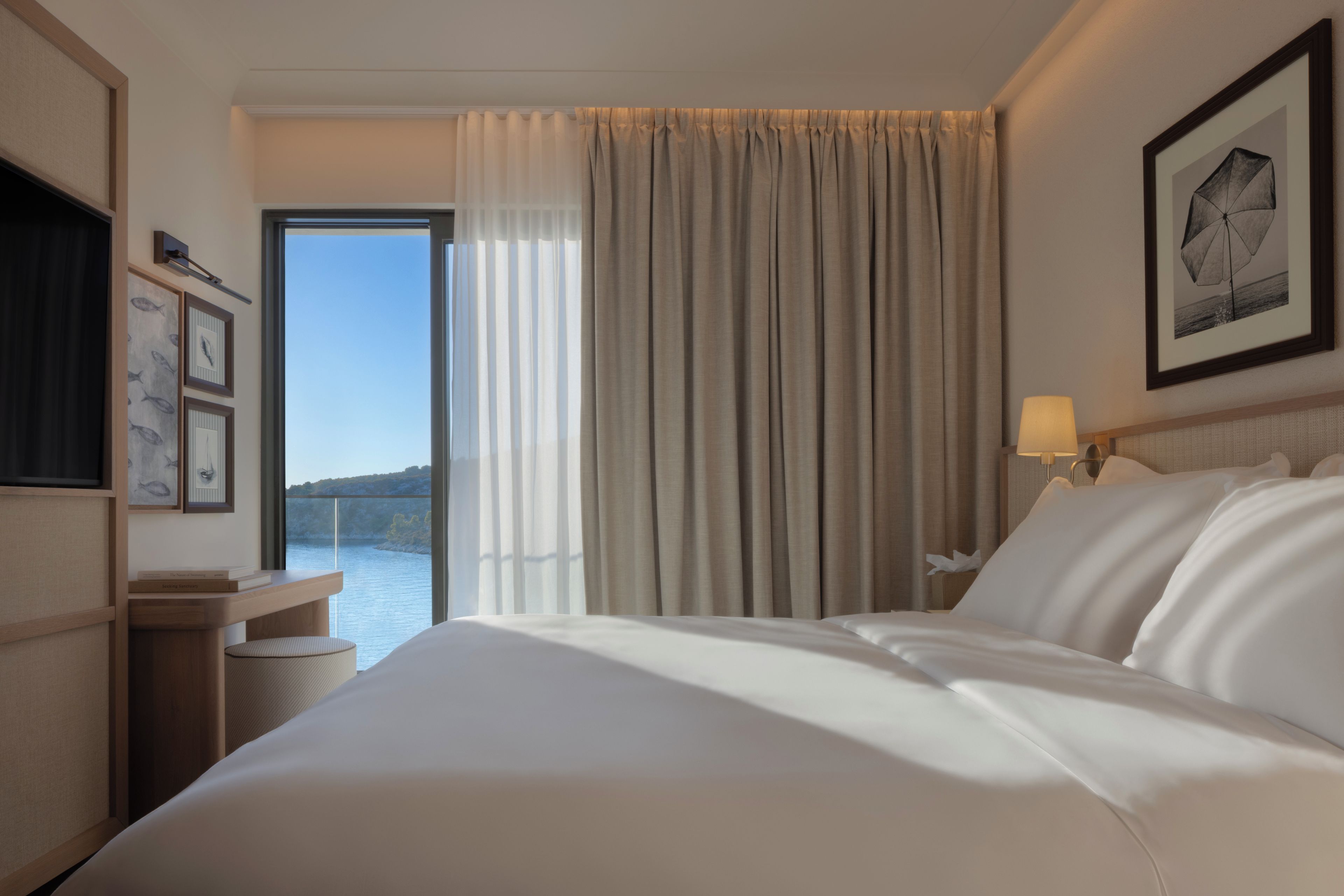 A hotel room with a bed, a bedside table, a lamp, a framed picture, and a view of the water.
