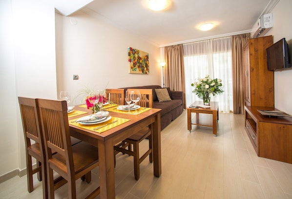 Un comedor con una mesa de madera preparada para cuatro personas, un sofá y una televisión al fondo.