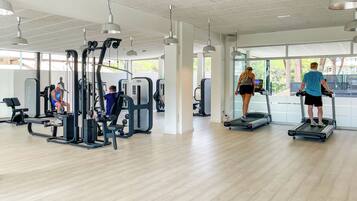 Un gimnasio con diversos equipos de ejercicio, incluyendo cintas de correr y máquinas de pesas, y ventanas amplias que permiten la entrada de luz natural.