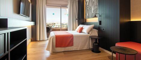 A hotel room with a bed, a TV mounted on the wall, a small table, and a view of the city through the window.