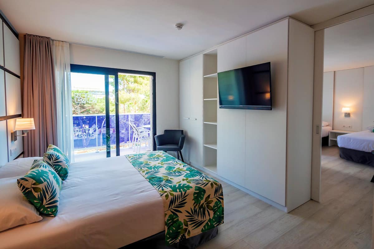 A hotel room with a bed, a TV mounted on the wall, a chair, and a balcony with a view of greenery.