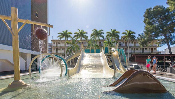 A water park with a water slide, splash pad, and palm trees. - Golden Port Salou & Spa (Salou)