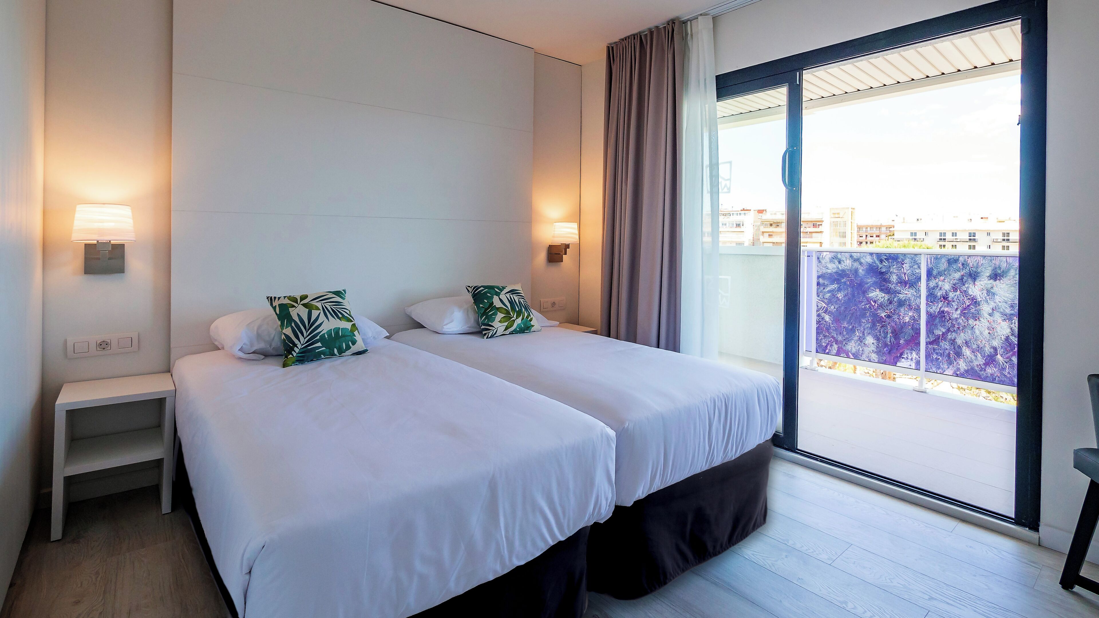 Habitación de hotel con dos camas, una mesita de noche, una lámpara y vista a la ciudad desde un balcón.