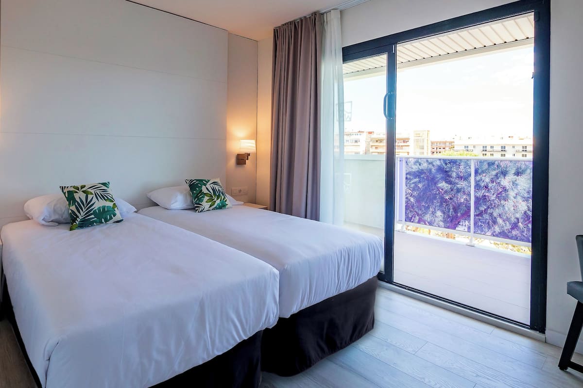 Habitación de hotel con dos camas, una mesita de noche, una lámpara y vista a la ciudad desde un balcón.