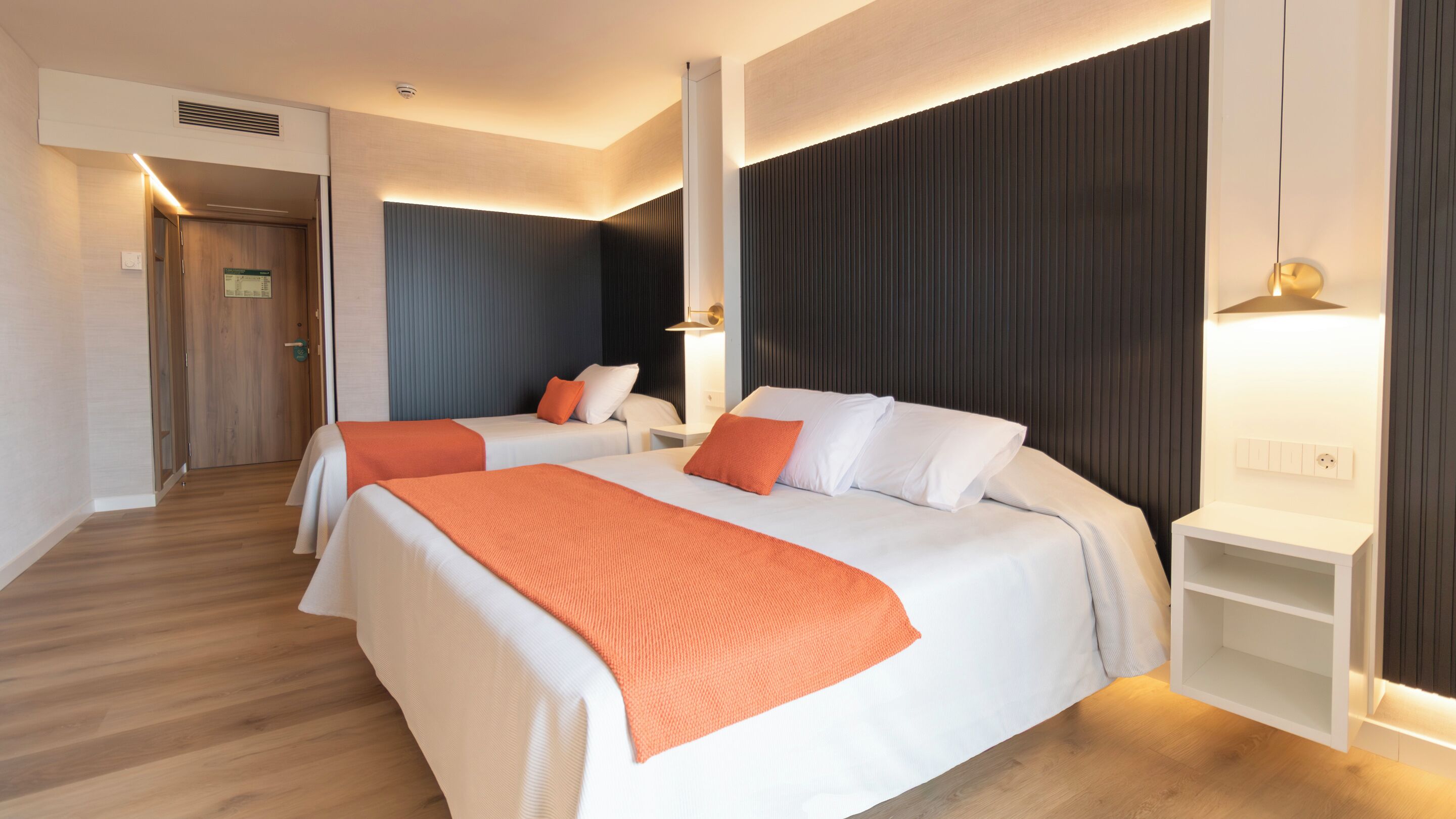A hotel room with two beds, a wooden floor, a dark accent wall, and a white nightstand.