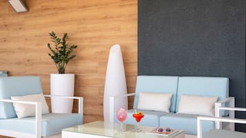 Una sala de estar moderna con un sofá azul claro, sillones blancos y una mesa de centro de vidrio. Un gran macetero blanco con follaje verde está a la izquierda, y una tabla de surf apoyada contra la pared.