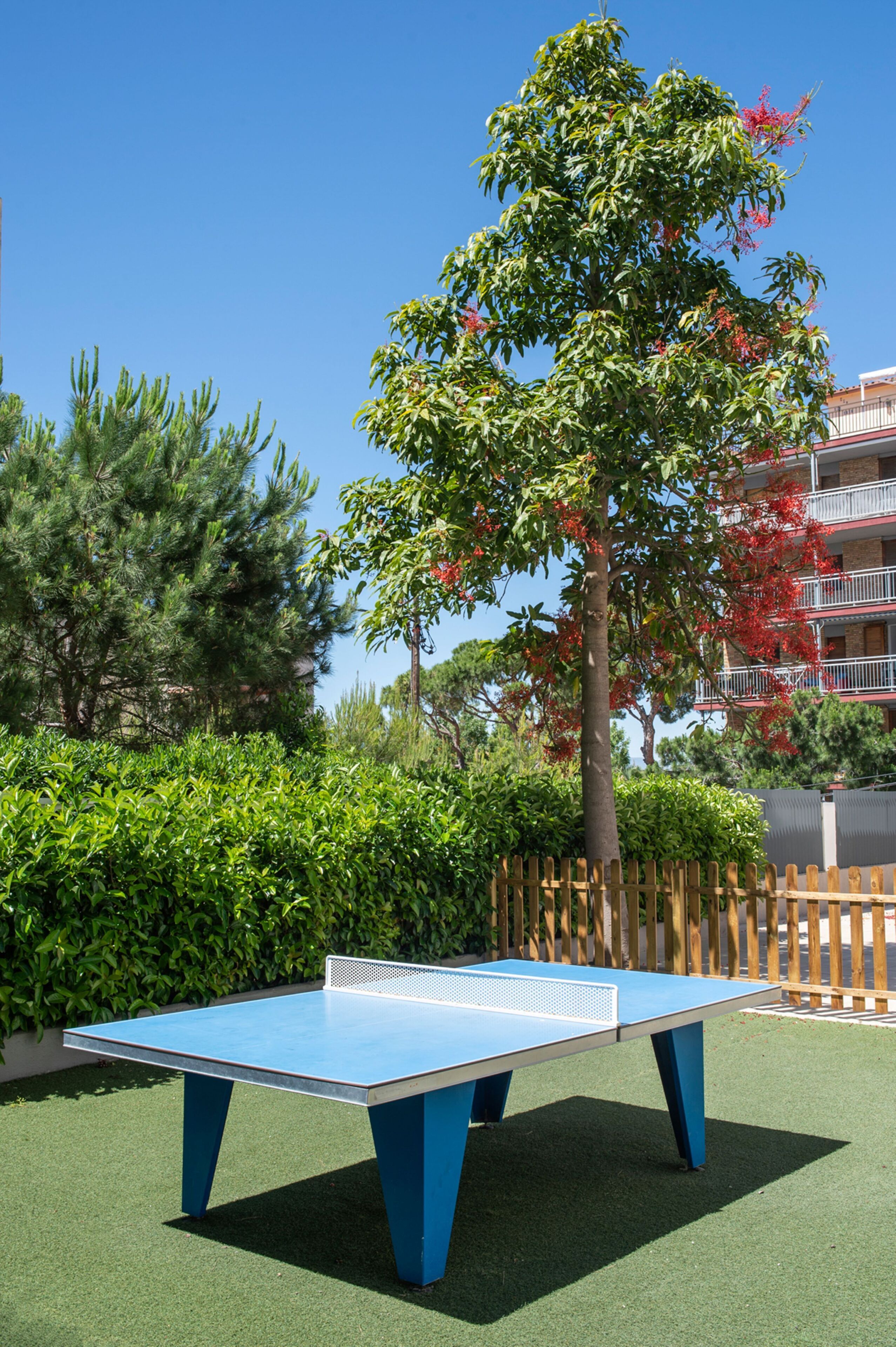 An outdoor ping pong table surrounded by lush greenery and a wooden fence.