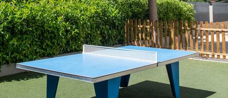An outdoor ping pong table surrounded by lush greenery and a wooden fence.
