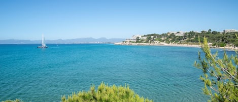 A coastal view with clear blue water, a sailboat, and a building on a hillside.