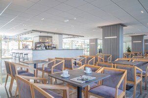 Un café moderno con mesas y sillas de madera, una zona de bar y grandes ventanales que permiten la entrada de luz natural.
