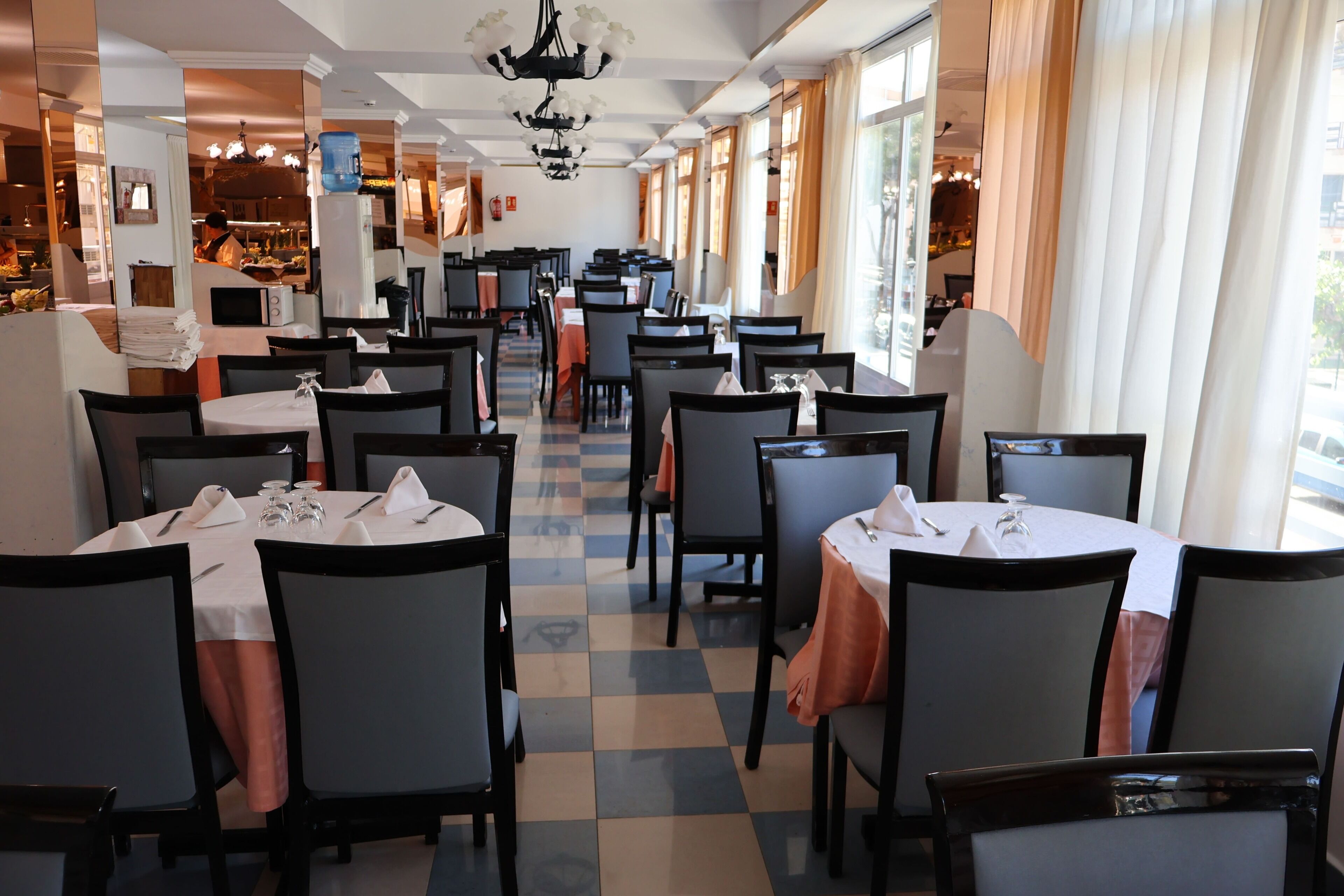 Un restaurante con varias mesas y sillas, suelo a cuadros y amplias ventanas con cortinas.