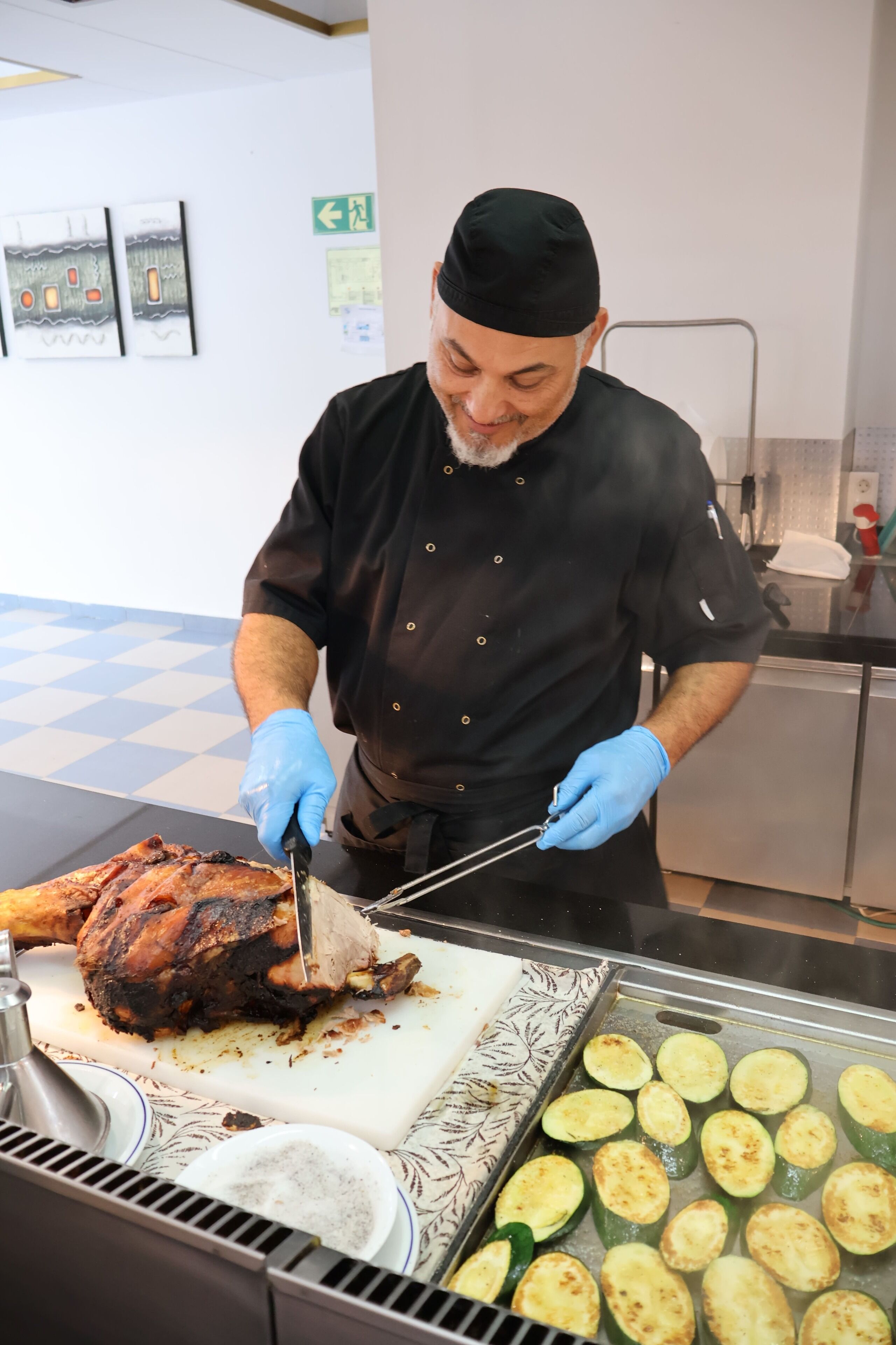 A chef carving a large roasted meat on a cutting board, with grilled vegetables on a tray nearby.