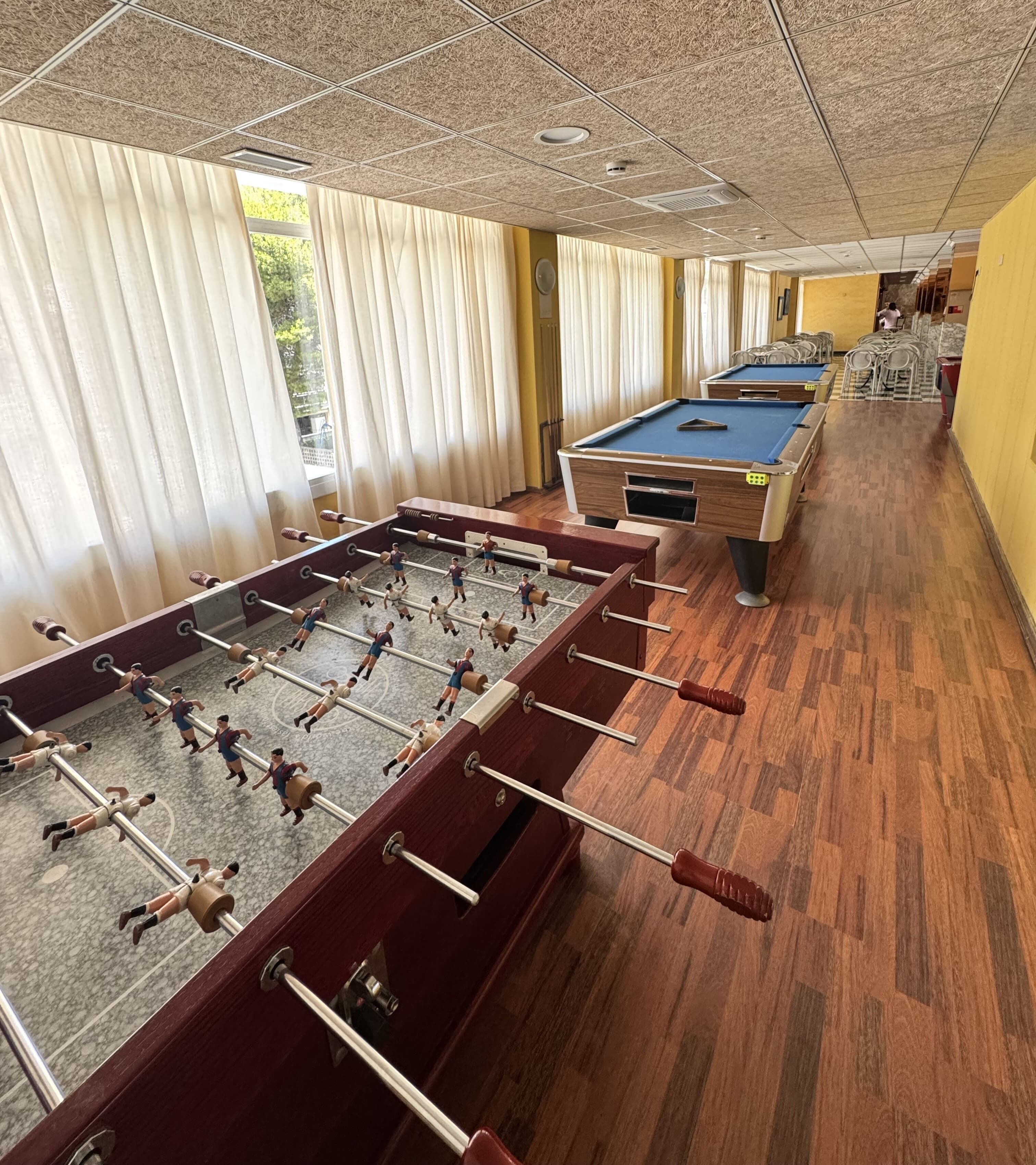 Mesa de billar y mesa de futbolín en una habitación con suelo de madera y ventanas amplias.
