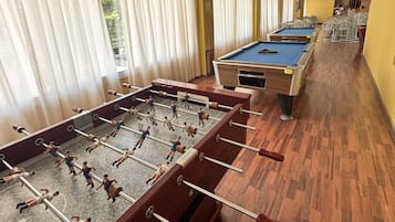 Mesa de billar y mesa de futbolín en una habitación con suelo de madera y ventanas amplias.