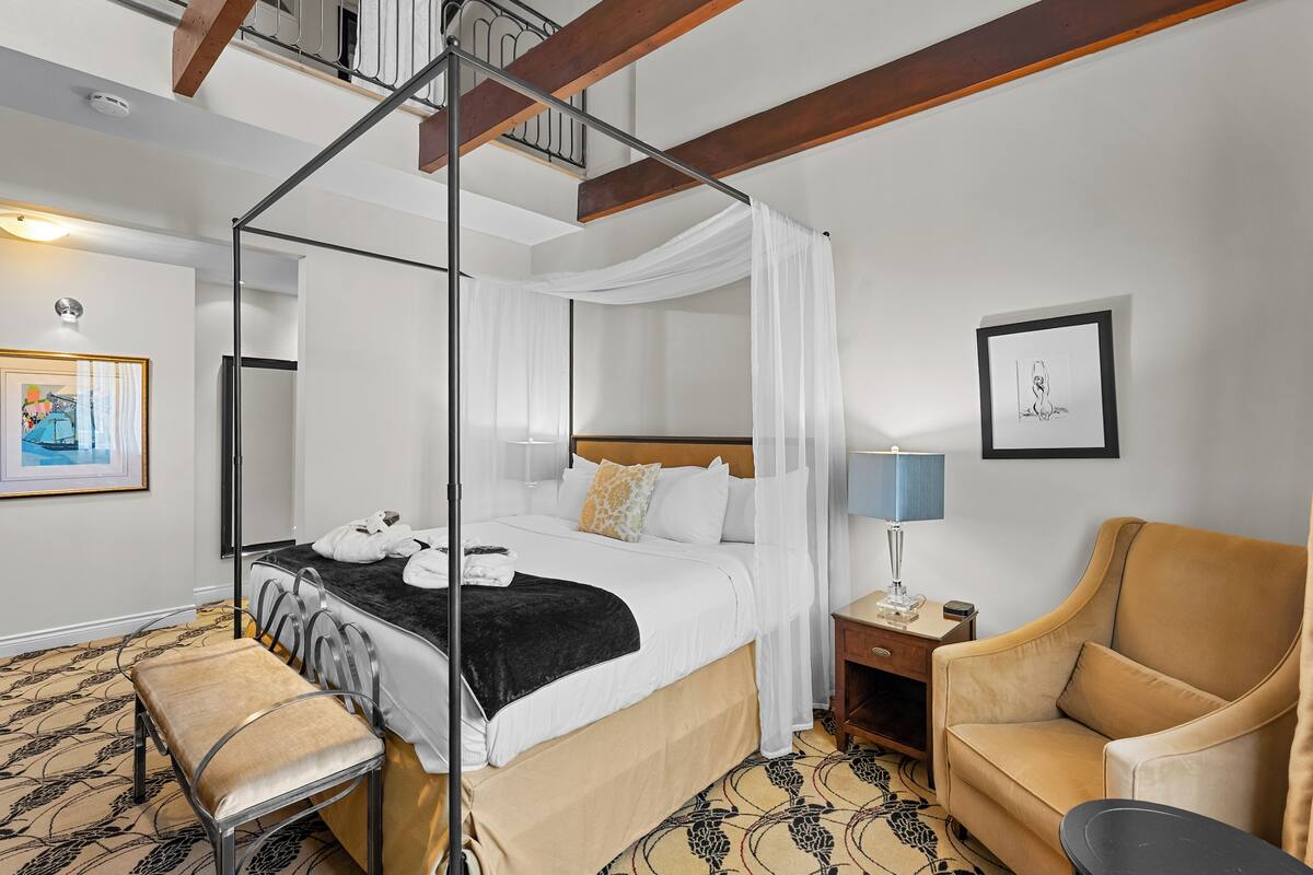 A hotel room with a four-poster bed, a bedside table with a lamp, a chair, and a bench.