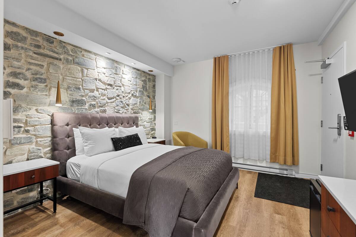 A modern bedroom with a stone wall, a tufted headboard, a wooden floor, a bed with a grey blanket, a yellow chair, and a TV on the wall.