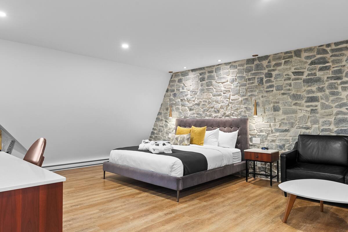 A modern bedroom with a stone accent wall, a bed with white bedding and a grey headboard, a wooden desk, a black leather chair, and a small table with a lamp.