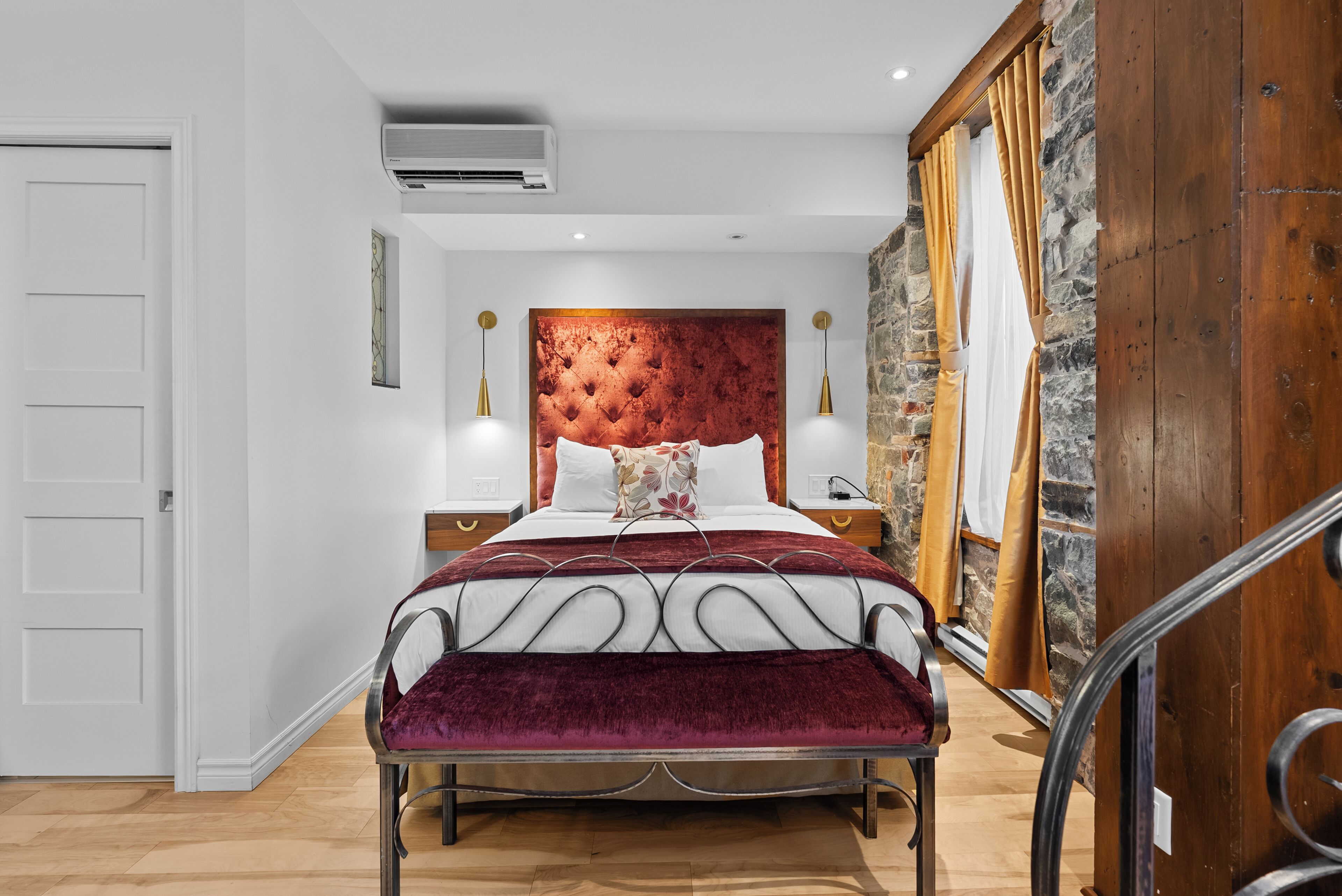 Un dormitorio con cabecera capitonada, una cama grande con colcha roja y blanca, un banco al pie de la cama y una pared de piedra a la derecha.