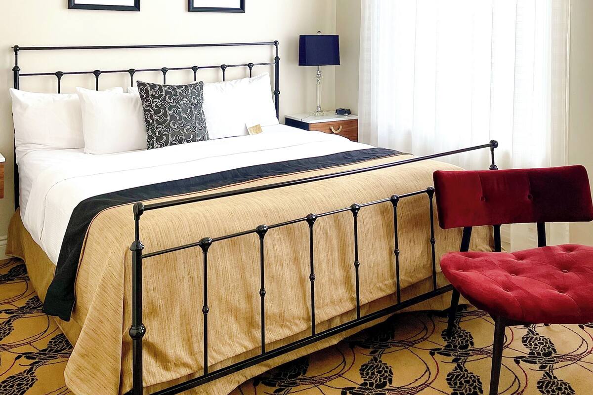 A neatly arranged hotel room with a bed, a bedside table with a lamp, and a window with sheer curtains.