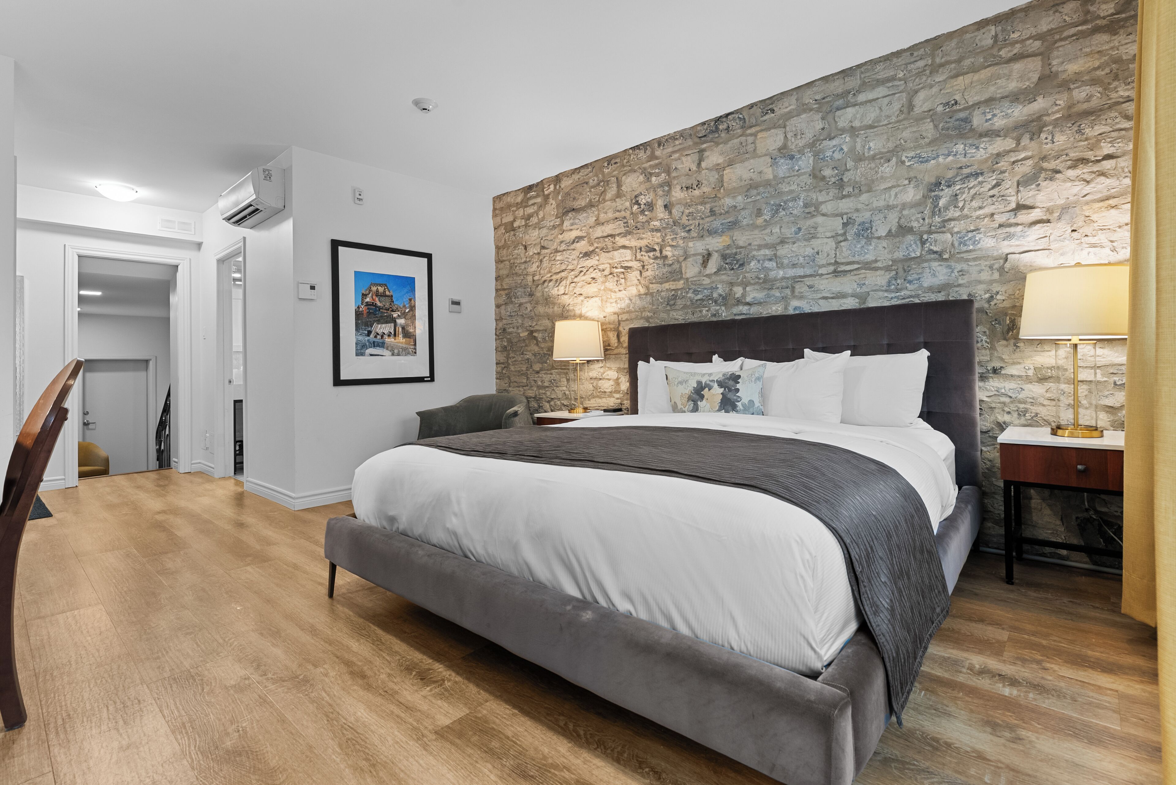 Un dormitorio con pared de piedra, una cama con ropa de cama blanca, un banco gris y suelo de madera.