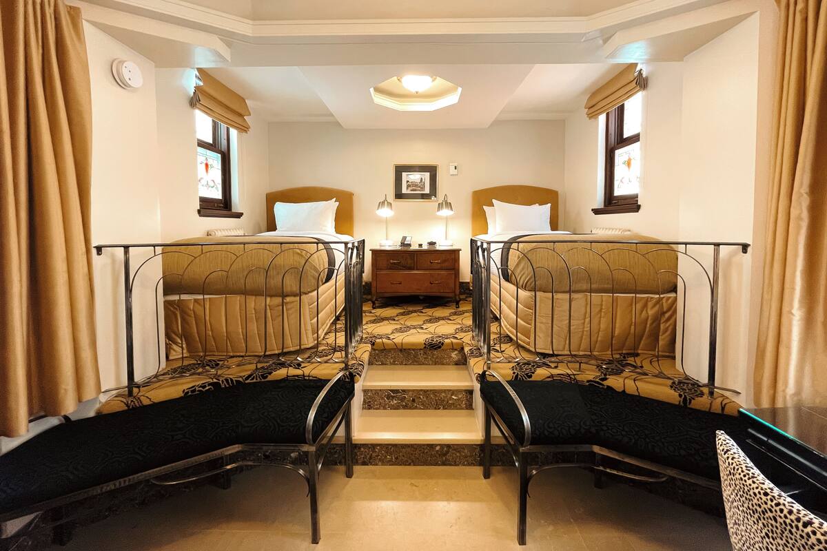 A hotel room with two beds, a nightstand, a lamp, and a window with curtains.