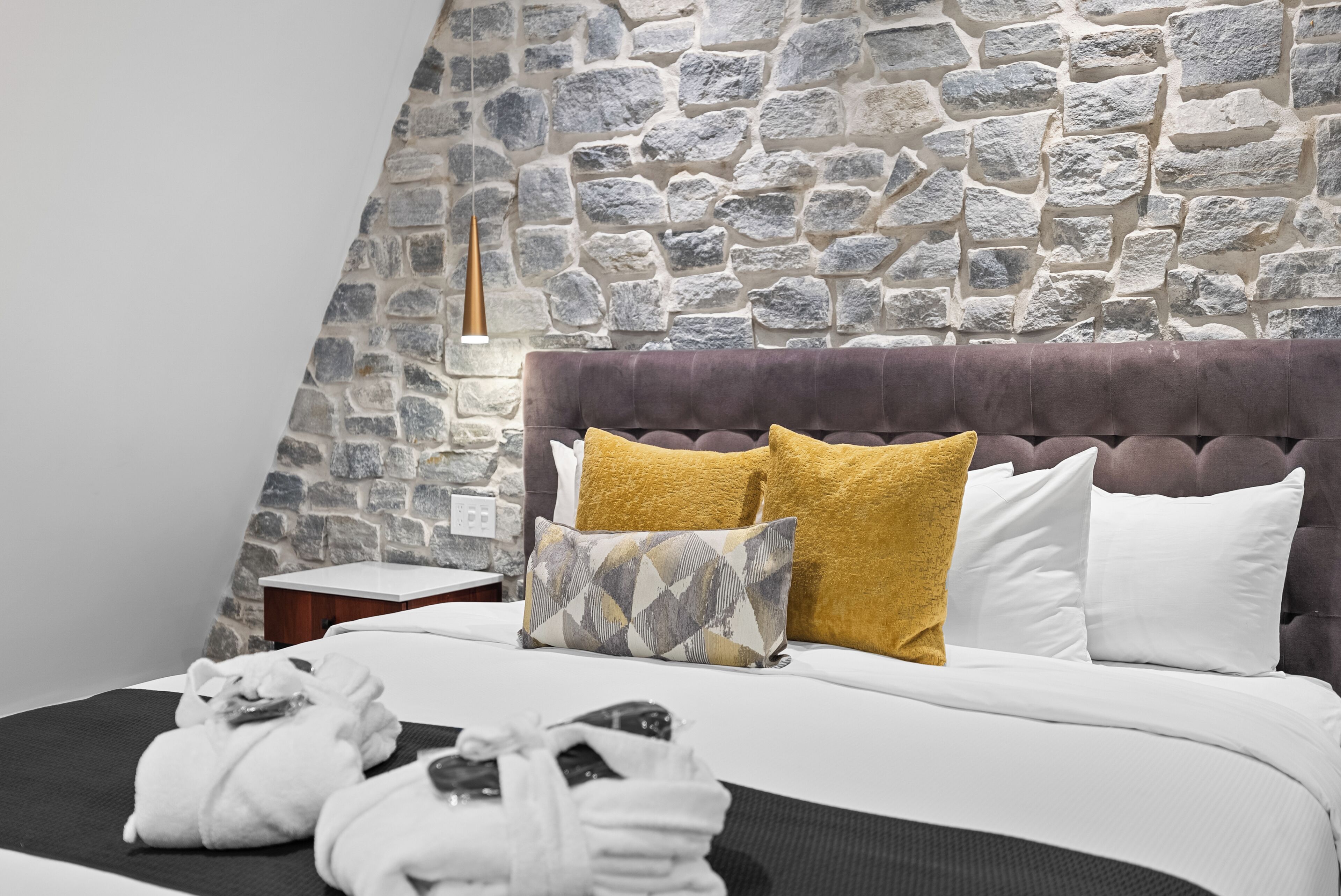Un dormitorio con pared de piedra, cabecera capitonada y mesita de noche con lámpara.