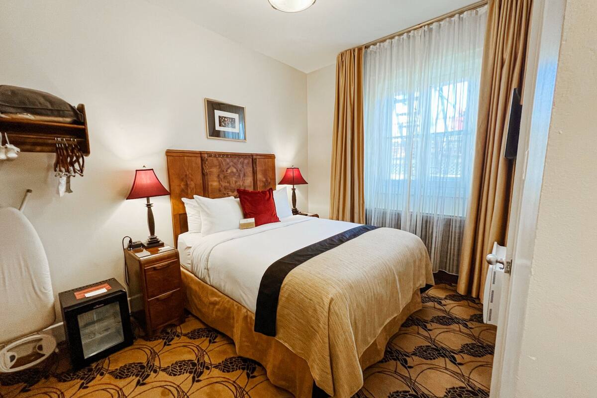 A hotel room with a large bed, two bedside lamps, a nightstand, a heater, and a window with curtains.