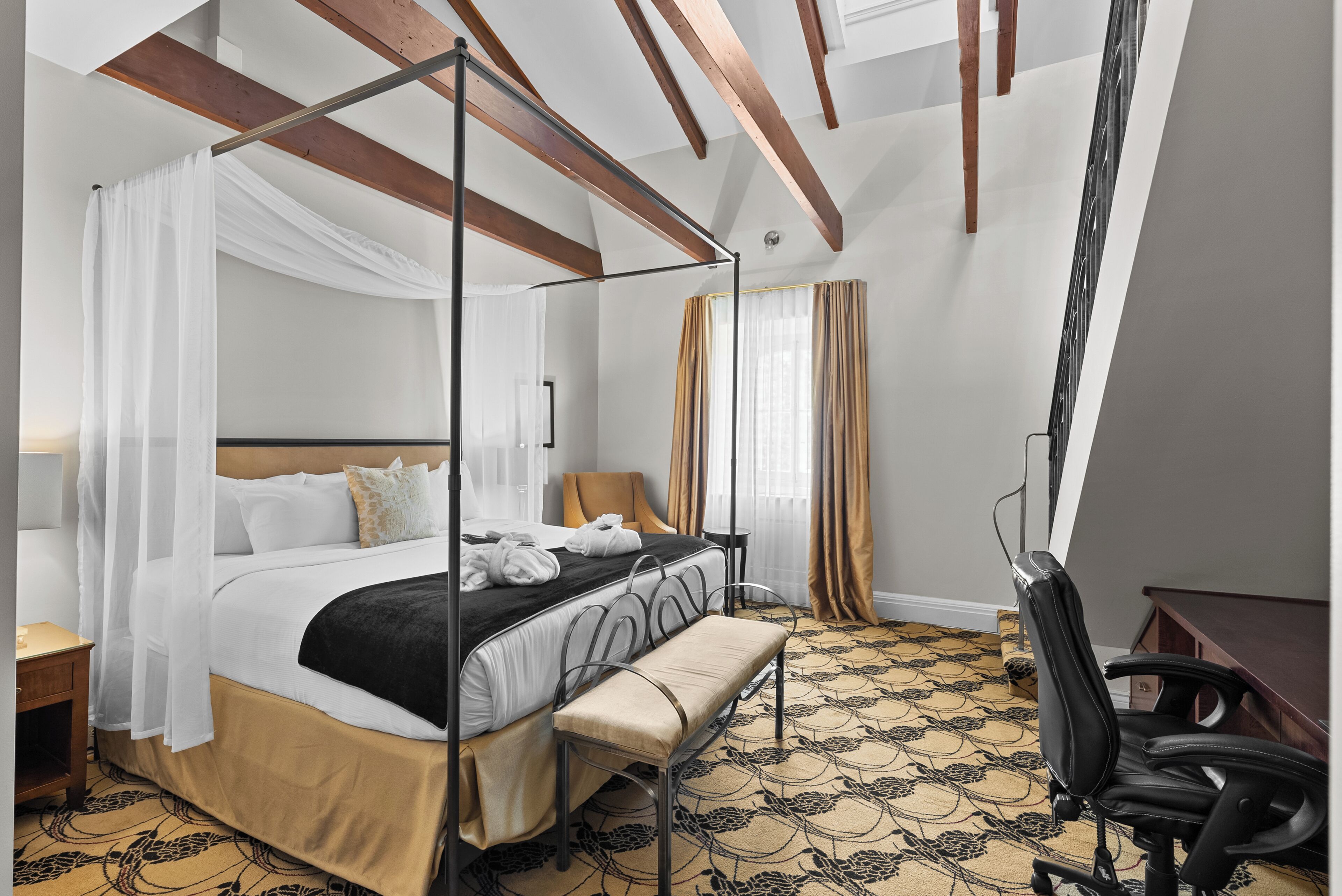 A four-poster bed with a canopy, a desk, and a chair in a hotel room with patterned carpet and wooden beams.