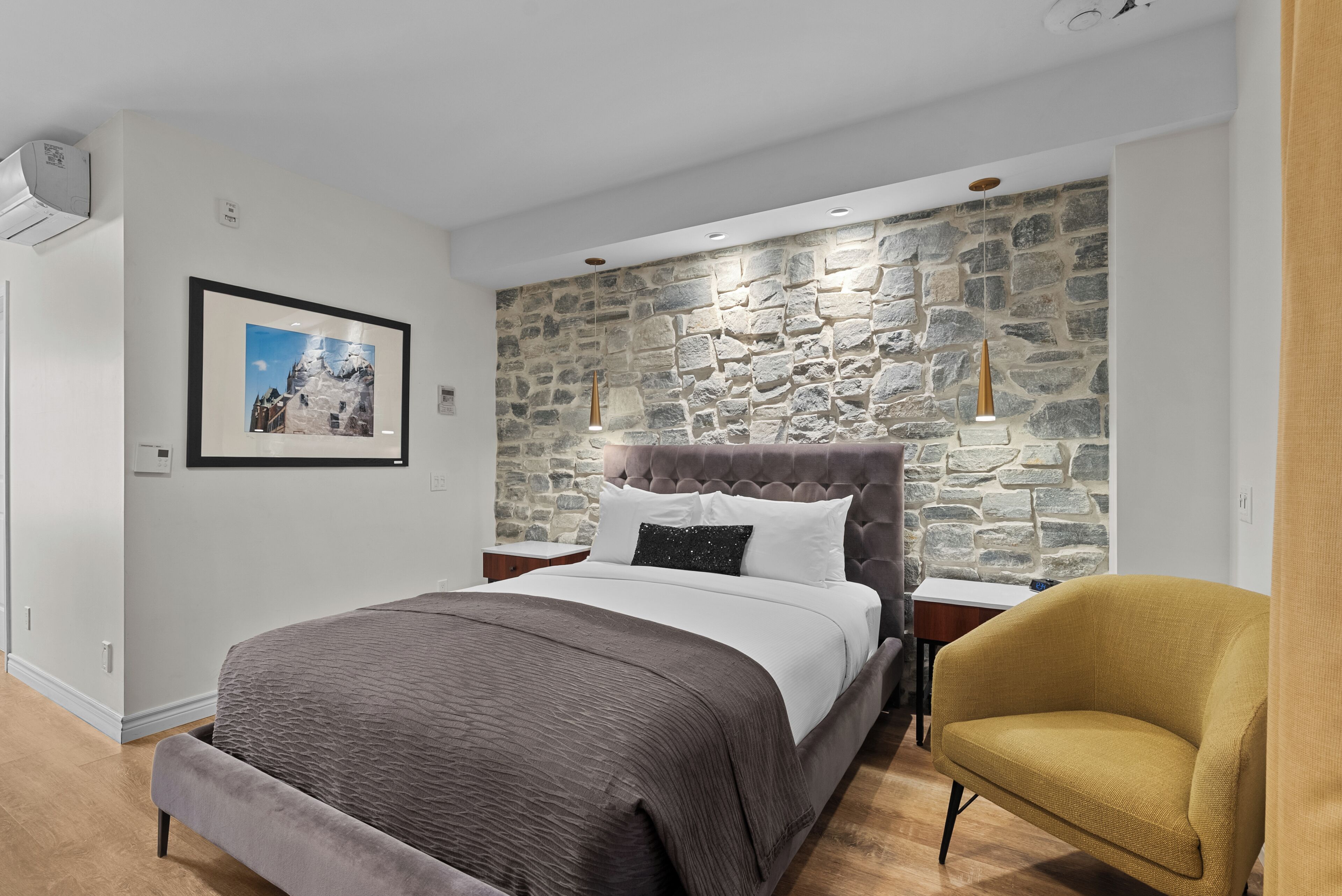 A modern bedroom with a stone wall, a large bed with a headboard, a bedside table, and a chair.