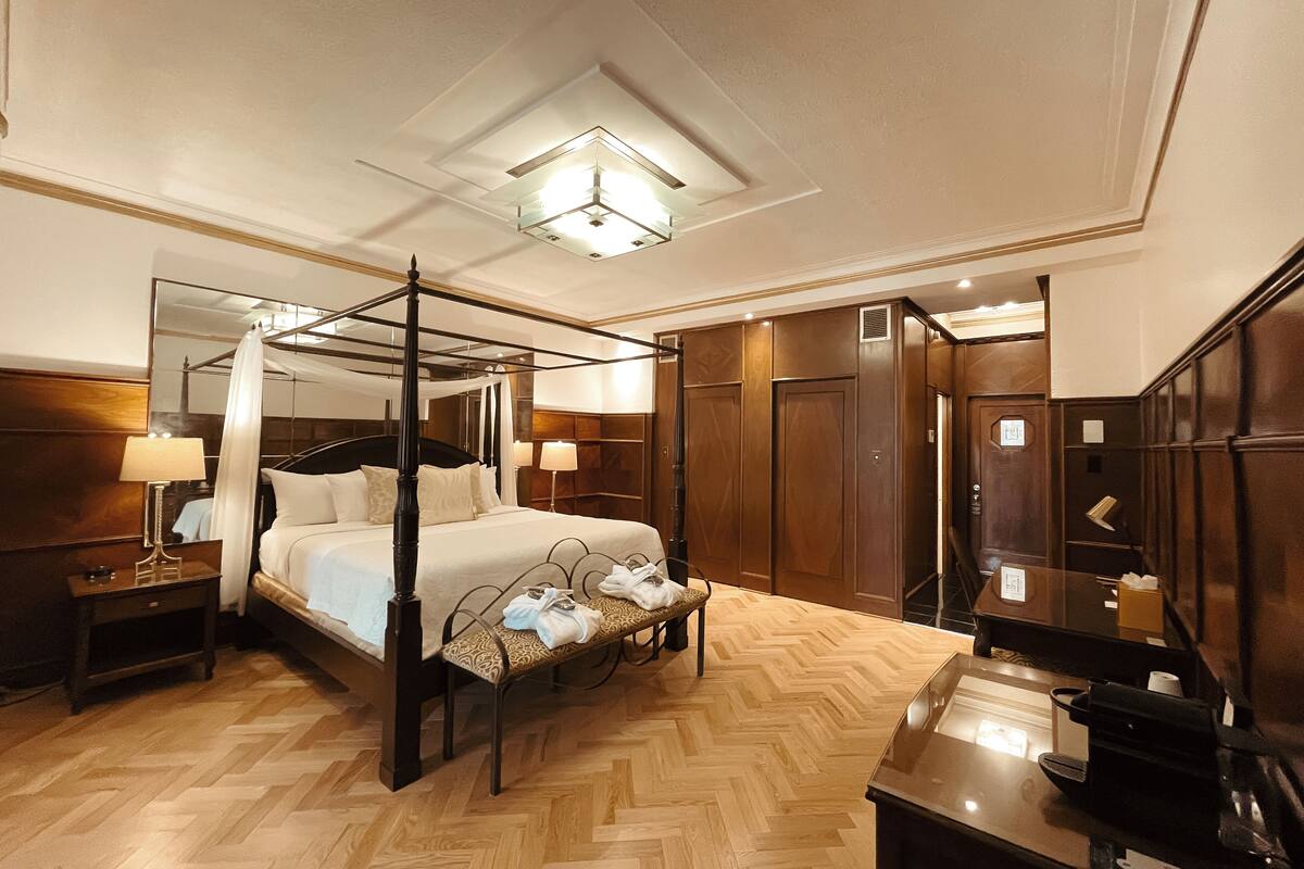 A four-poster bed with a canopy, a wooden floor, a desk with a lamp, and a mirror.