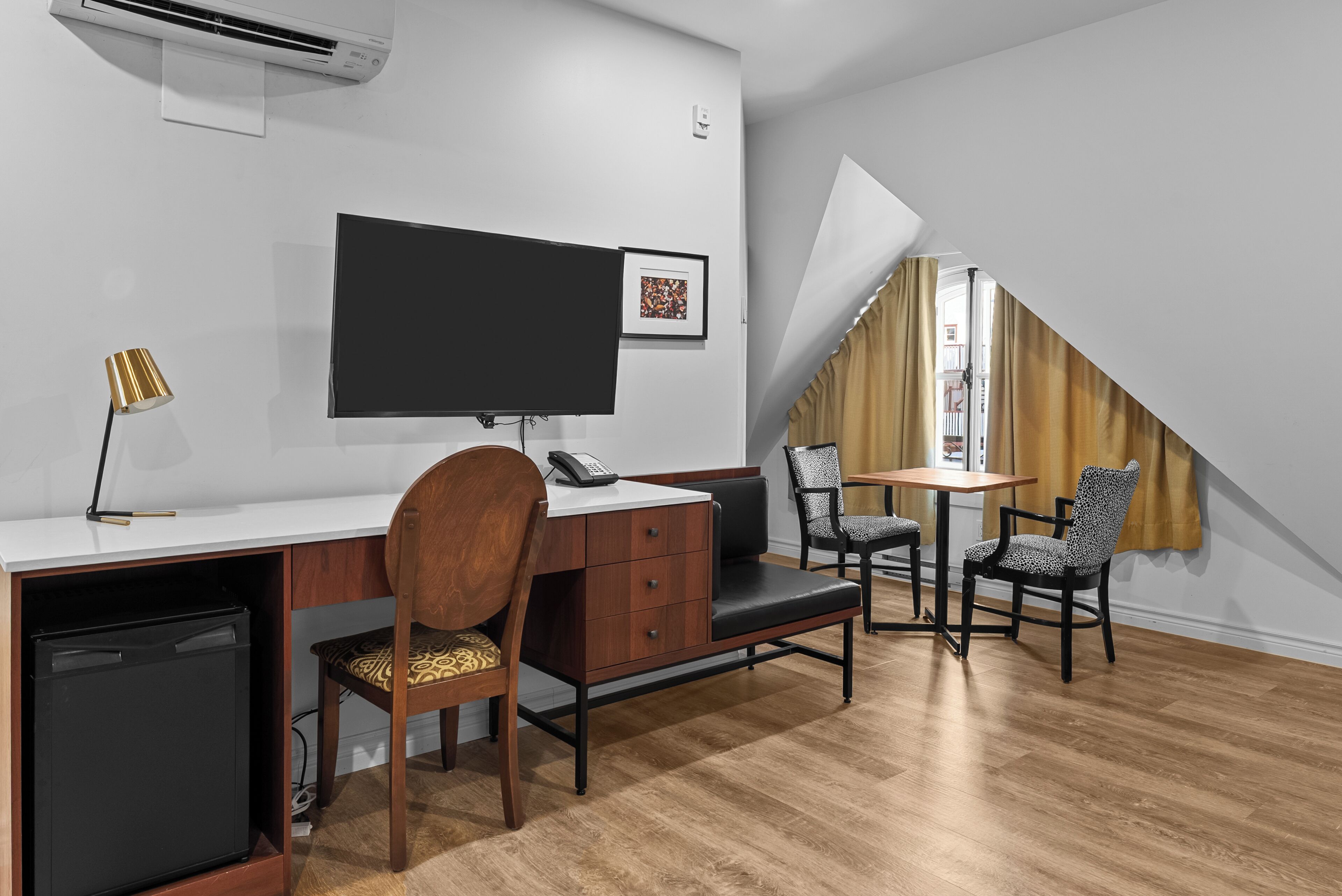 Una habitación de hotel compacta con escritorio, una silla, un refrigerador pequeño, un televisor y una ventana con cortinas.